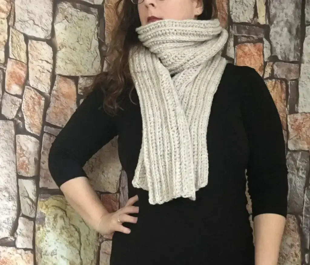 A woman stands against a backdrop of a stone facade, wearing a black shirt and a white crocheted scarf.